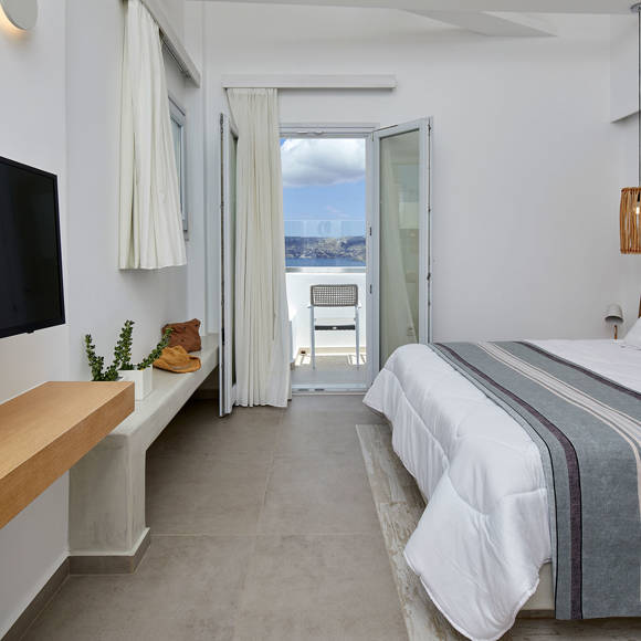 Santorini View Hotel double bed with white and grey linen, wooden decoration, white curtains and balcony with sea view